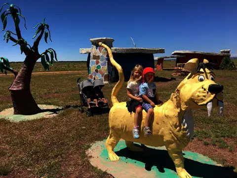 Two children sit on a yellow saber-toothed cat statue outdoors.
