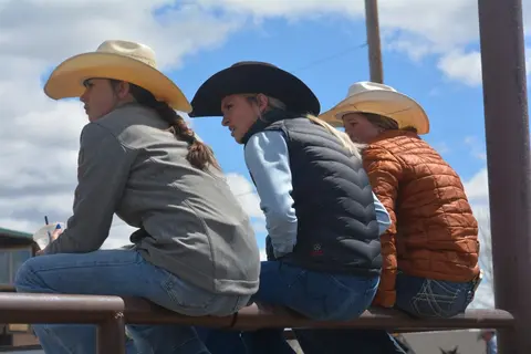 Three young women wearing cowboy hats sit on a rail watching an event.
