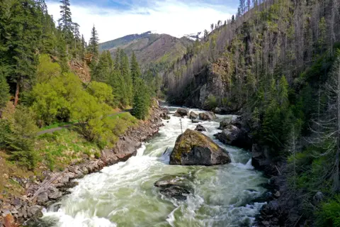 A river flows through a rocky canyon with trees and mountains.