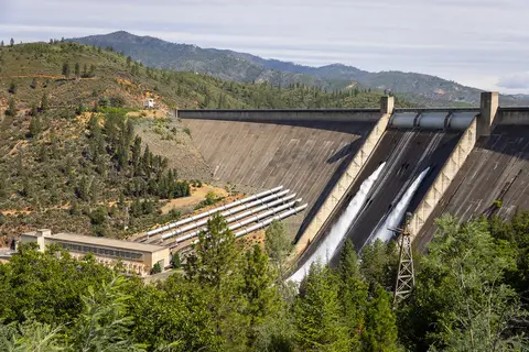 A large concrete dam with water rushing down its spillways and mountains in the background.