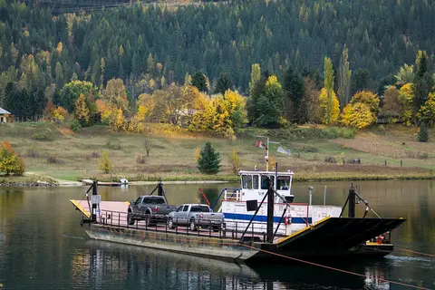 A car ferry crossing a body of water with a forested hill in the background.