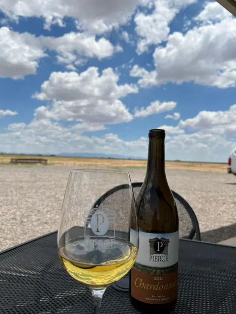 A glass and a bottle of wine sit on a table outside against a cloudy sky.