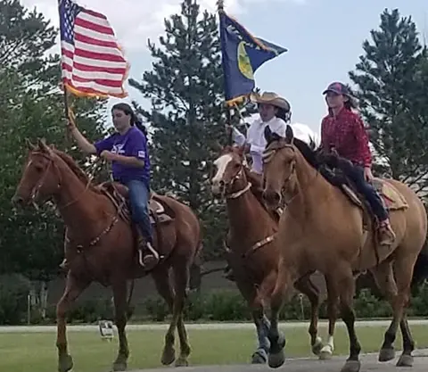 Three people ride horses while carrying flags outdoors.