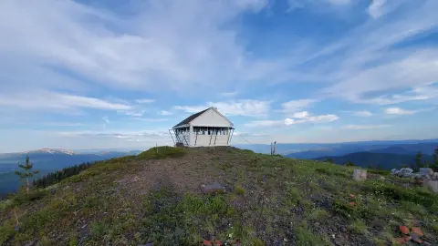 A small white building sits on top of a mountain under a cloudy blue sky.