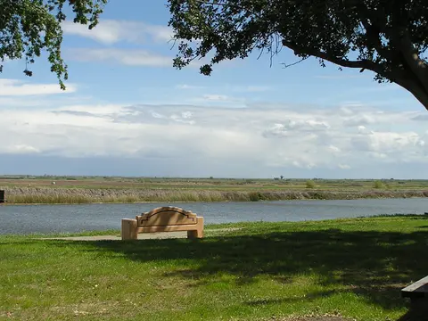 A bench sits near a river with fields in the background under a cloudy sky.