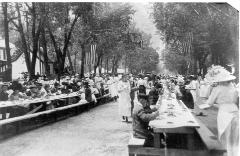 A historic black and white photo shows a large outdoor gathering at long tables under trees decorated with American flags.