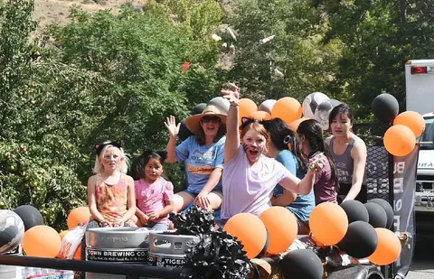A group of people ride on a float decorated with balloons and kegs during an outdoor event.