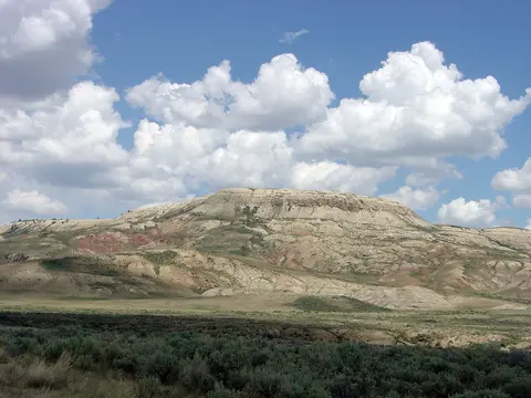 A landscape showing a large barren hill under a blue sky with white clouds.