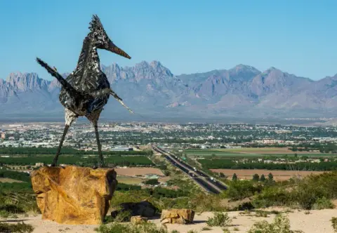 A large metal roadrunner sculpture overlooks a valley with mountains in the background.