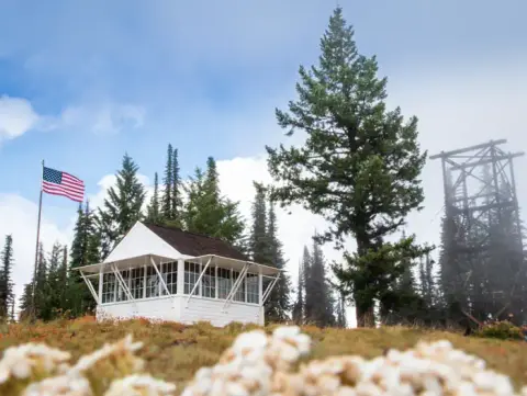 A white cabin and an American flag stand on a mountain ridge among trees.