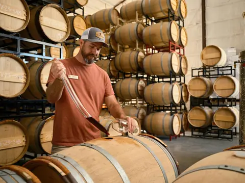 A man uses a wine thief to sample wine from a wooden barrel inside a cellar.