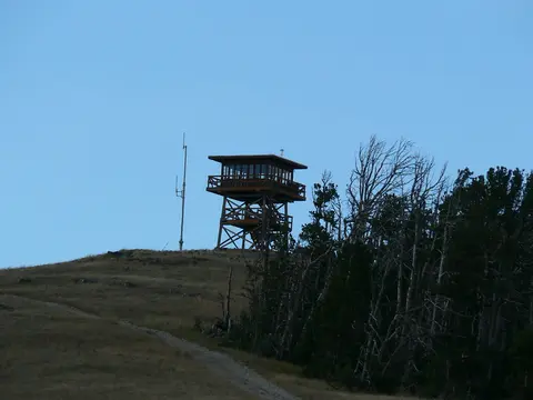 A fire lookout tower stands on top of a hill next to trees under a blue sky.