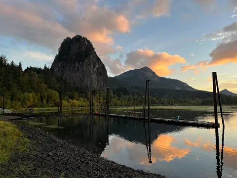 A wooden dock extends into a calm body of water with a rocky mountain rising in the background at sunset.