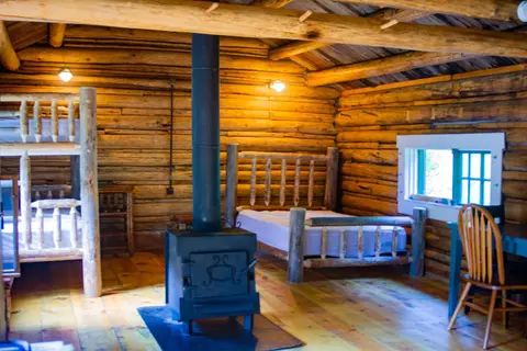 The interior of a rustic log cabin with beds and a wood stove.