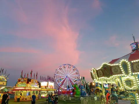 A brightly lit carnival rides and booths stand under a colorful sunset sky.