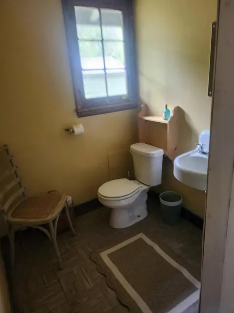 A small bathroom with a toilet, sink, window, and a chair.