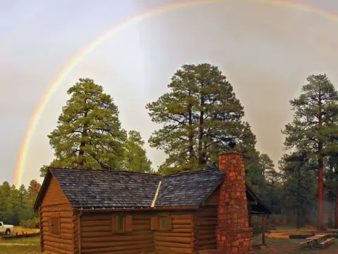 A log cabin sits among tall pine trees beneath a rainbow in the sky.