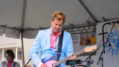 A man in a light blue suit plays a guitar under a tent.