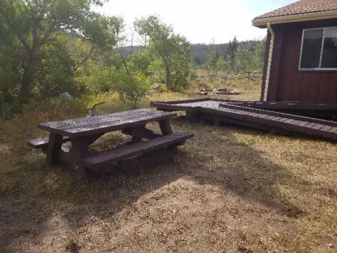 A wooden picnic table sits on dry grass outside a brown building with a deck.