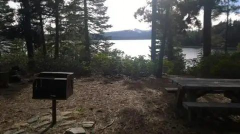 A campsite with a picnic table and grill overlooking a lake through trees.