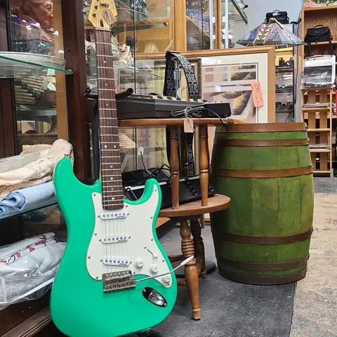 A bright green electric guitar stands inside a cluttered shop near a green wooden barrel.