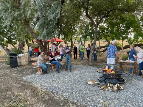 A group of people gather outdoors under trees near a fire pit.