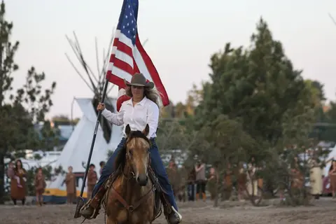 A woman in a cowboy hat rides a horse carrying an American flag.