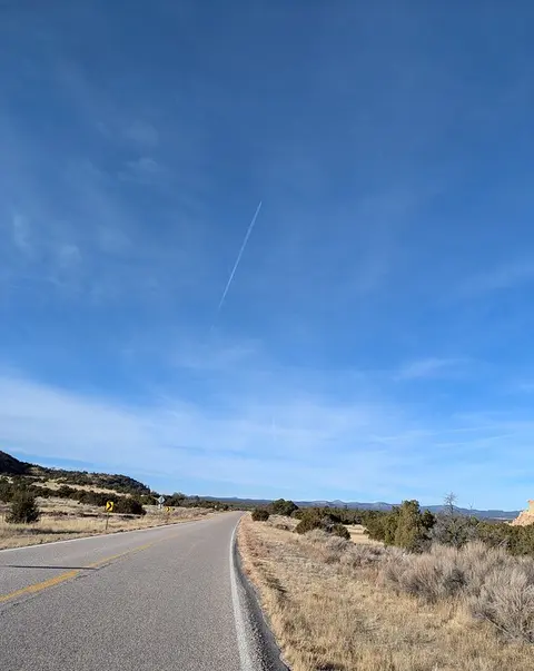 A paved road winds through an arid landscape under a blue sky with a contrail.