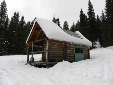 A snow-covered log cabin sits in a snowy landscape surrounded by pine trees.