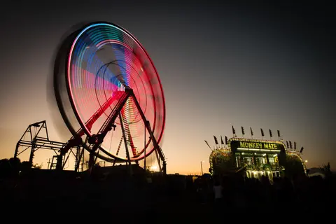 A long-exposure photograph of a spinning Ferris wheel and a lit carnival attraction at dusk.