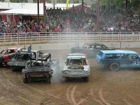 Several damaged cars compete in a demolition derby before a large crowd of spectators.
