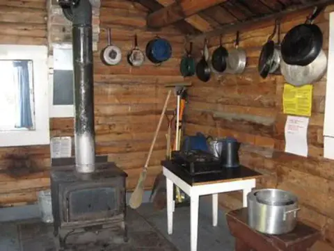 An interior view of a rustic log cabin with a wood-burning stove and hanging cookware.