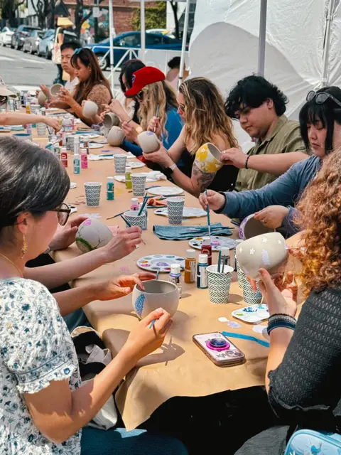 People sit at a long table outdoors and paint pottery bowls.