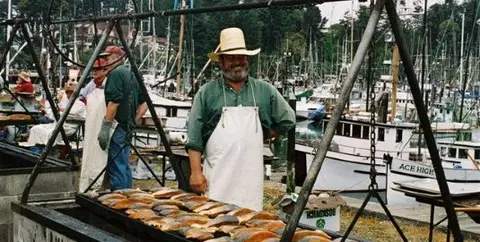 A man stands near an outdoor fish grill at a harbor with boats.