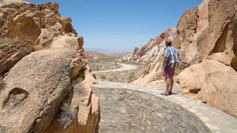 A person stands on a stone walkway overlooking a desert landscape with a winding road.