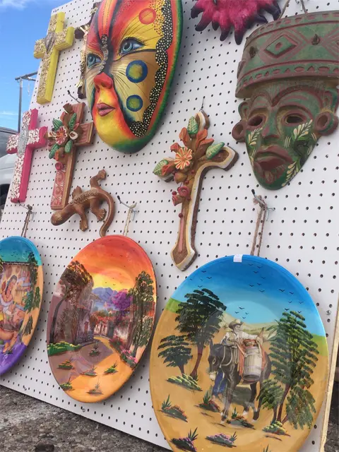 Various handcrafted decorative items, including masks, crosses, and painted plates, are displayed on a pegboard.