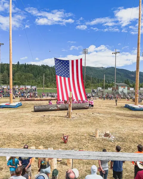 A giant American flag is suspended between two tall wooden poles at an outdoor arena with logs and spectators.