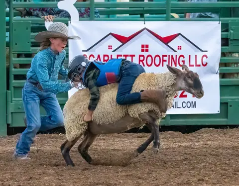 A child rides a sheep in a rodeo arena while another person runs beside them.