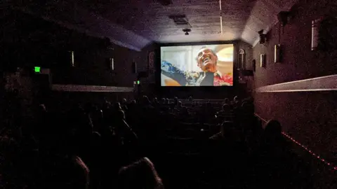 An interior view of a dark movie theater with an audience watching a film on screen.
