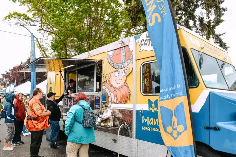 People line up to order food from a colorful food truck at an outdoor event.