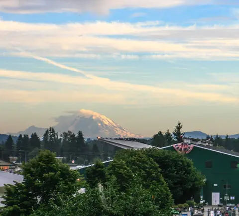 A scenic view of a snow-capped mountain behind trees and buildings under a cloudy sky.