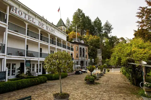 A large historic white hotel with multiple balconies stands beside a brick courtyard and wooded hillside.