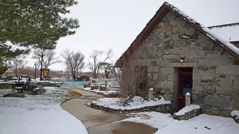 A stone building with a snow-covered roof stands near a snowy path and swimming pool.