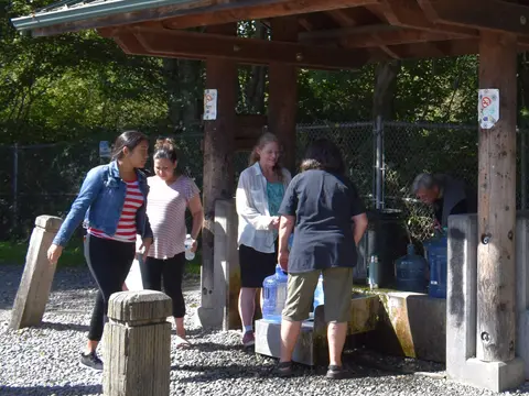People gather around a water filling station under a wooden shelter outdoors.