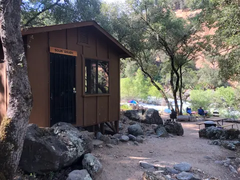 A small brown cabin with a sign that reads SOUR DOUGH stands near a river in a wooded area.