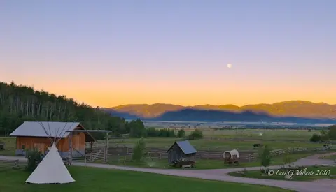 A scenic landscape showing a rustic ranch at dusk with mountains and a moon in the sky.