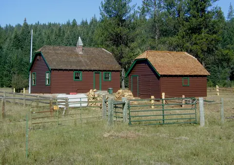 Two brown wooden buildings with green trim stand in a grassy field near a forest.