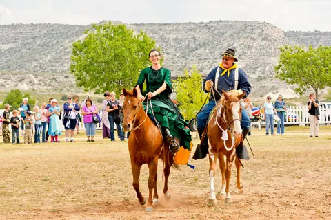 A woman in a green dress and a man in costume ride horses outdoors with spectators in the background.