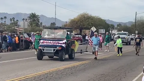 A golf cart decorated for a parade is driven down a street with people walking alongside.