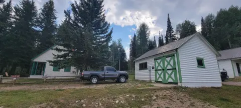 A pickup truck parked between two white log buildings surrounded by trees under a cloudy sky.
